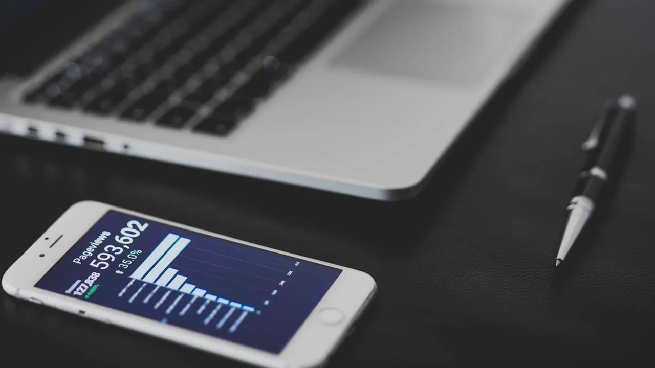 Phone with a revenue dashboard beside a laptop and pen